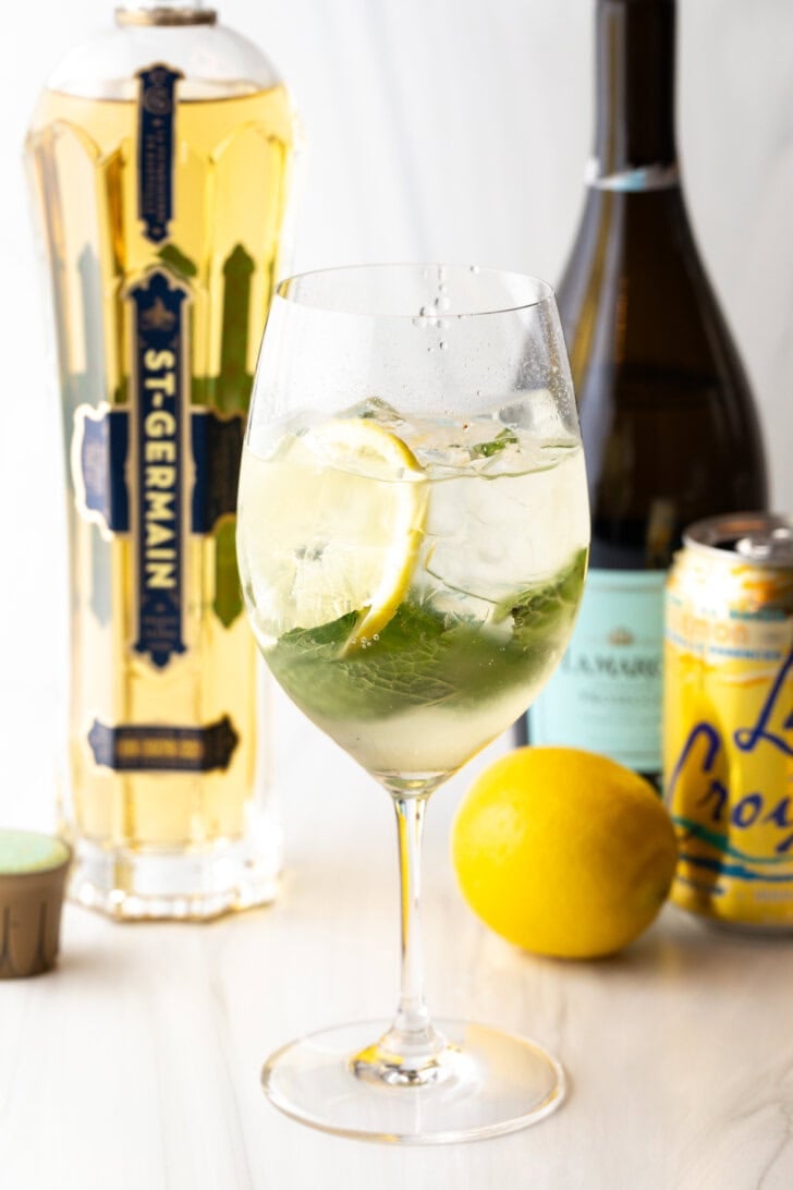 Wine glass with alcohol, mint leaves, and lemon slices. In the background of the hugo spritz is a whole lemon and bottles of prosecco, st. germain liquor, and a can of la croix soda.