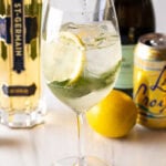 Wine glass with alcohol, mint leaves, and lemon slices. In the background are a whole lemon and bottles of prosecco, st. germain liquor, and a can of la croix soda.