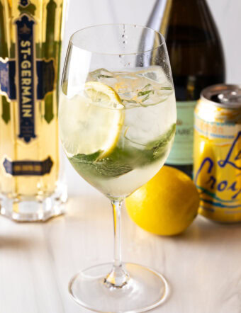 Wine glass with alcohol, mint leaves, and lemon slices. In the background are a whole lemon and bottles of prosecco, st. germain liquor, and a can of la croix soda.