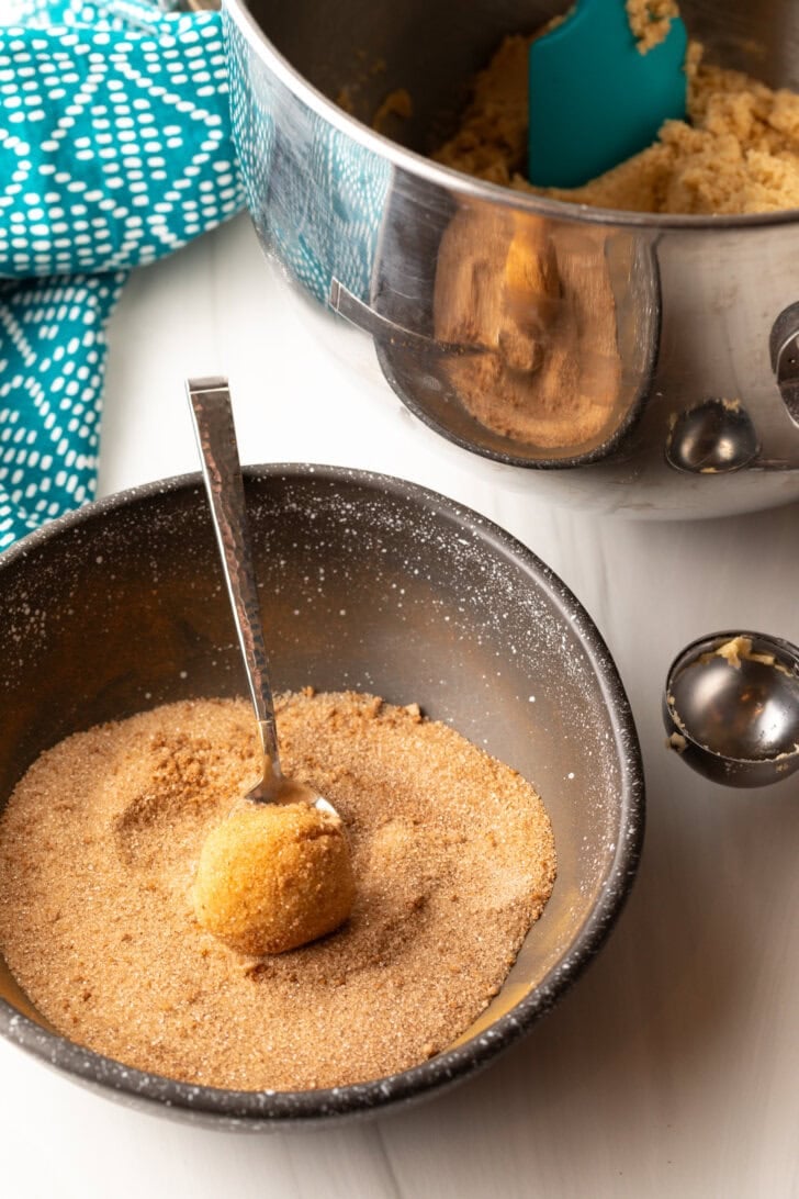A metal spoon rolling a cookie dough ball around in a bowl of cinnamon and sugar.