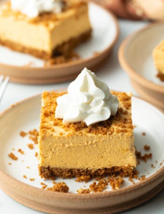 Square of pumpkin pie cheesecake on a white plate, topped with whipped cream.