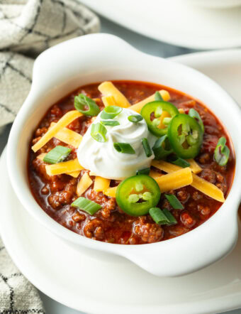 Top down view of a white bowl of beanless chili topped with shredded cheese, jalapeno slices, sour cream and chopped green onions.