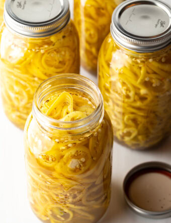 4 large jars of canned pickled banana peppers. The jar closest to the camera has the lid off showing the slices of peppers filling the jar.