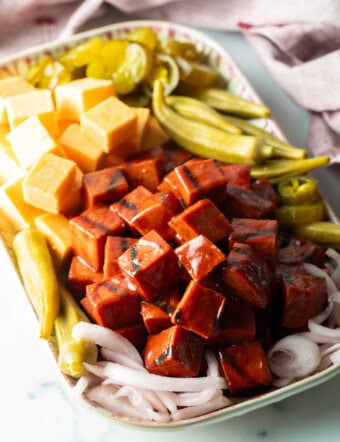 Serving plate with a stack of bbq smoked bologna cubes, squares of cheese, pickled okra, slices of red onion, and pickles.