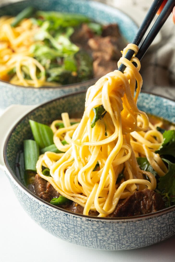 Hand with chopsticks grabbing noodles from the bowl of soup with beef.