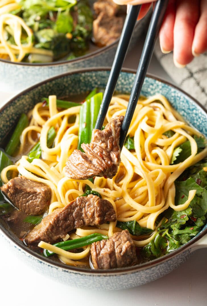 Hand with chopsticks grabbing a piece of meat from the bowl of soup with noodles.