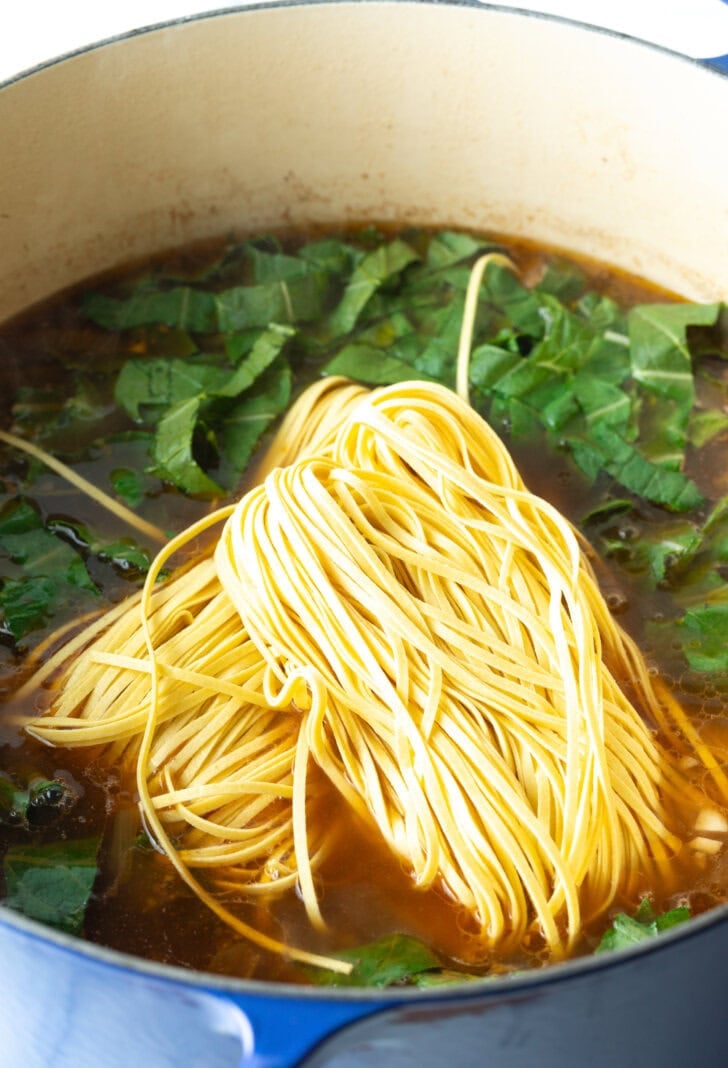Adding long pasta noodles to the pot of cooking beef broth and greens.