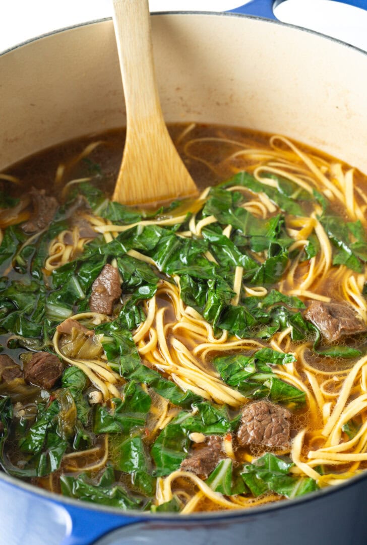 Top down view large pot of soup with noodles and greens, stirred by a wooden spatula.