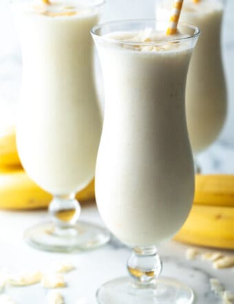 3 large mix drink glasses with banana smoothie and a yellow striped straw in each glass.