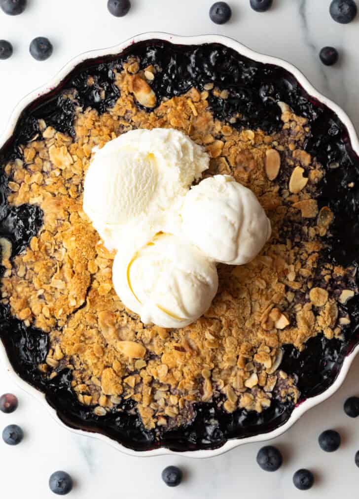 Scalloped round baking dish with blueberry crisp, topped with scoops of ice cream.