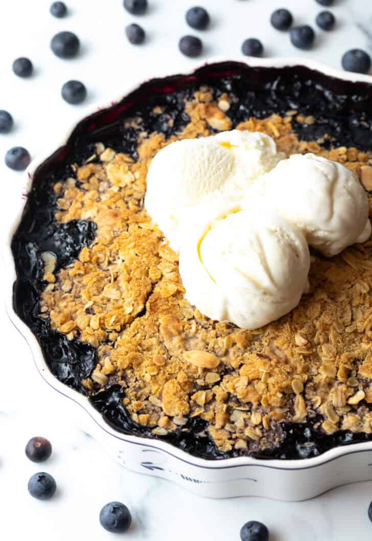 Scalloped round baking dish with blueberry crisp, topped with scoops of ice cream.