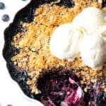 Scalloped round baking dish with blueberry crisp, topped with scoops of ice cream. A metal spoon has taken away a serving of the crumble.