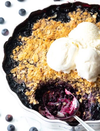 Scalloped round baking dish with blueberry crisp, topped with scoops of ice cream. A metal spoon has taken away a serving of the crumble.