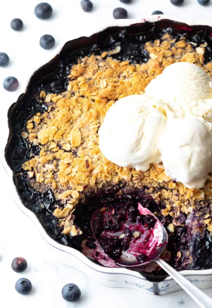 Scalloped round baking dish with blueberry crisp, topped with scoops of ice cream. A metal spoon has taken away a serving of the crumble.