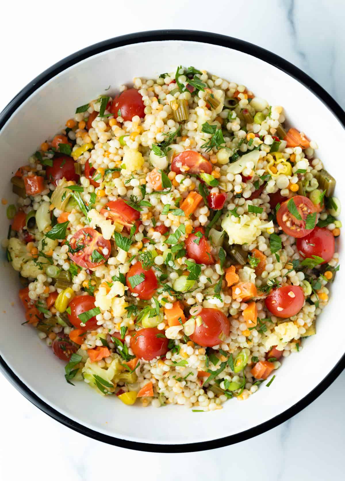 Top down view pearl couscous salad with chopped tomatoes, fresh herbs, and pickled vegetables in a large white mixing bowl.