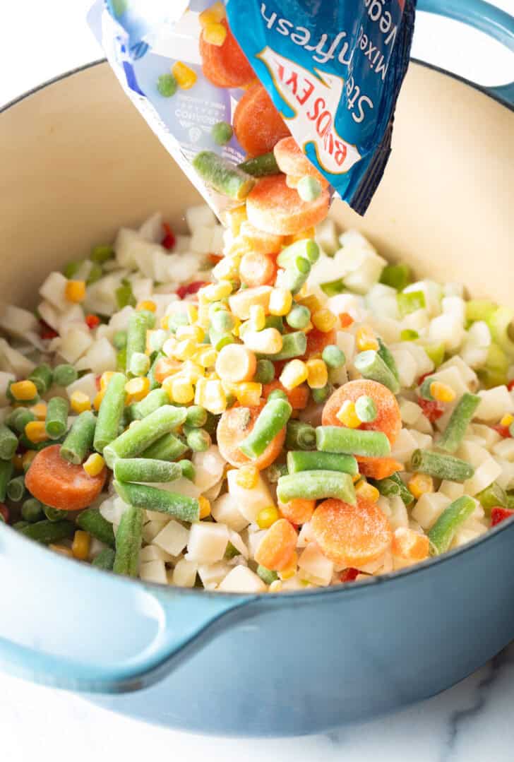 Pouring a mix of frozen corn, green beans, carrots, and peas into a large cooking pot.