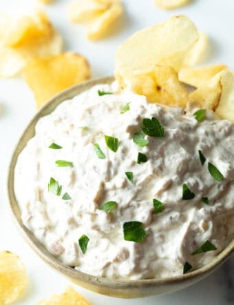 Bowl of creamy white French onion dip, topped with chopped chives and with a potato chip stuck into the dip.