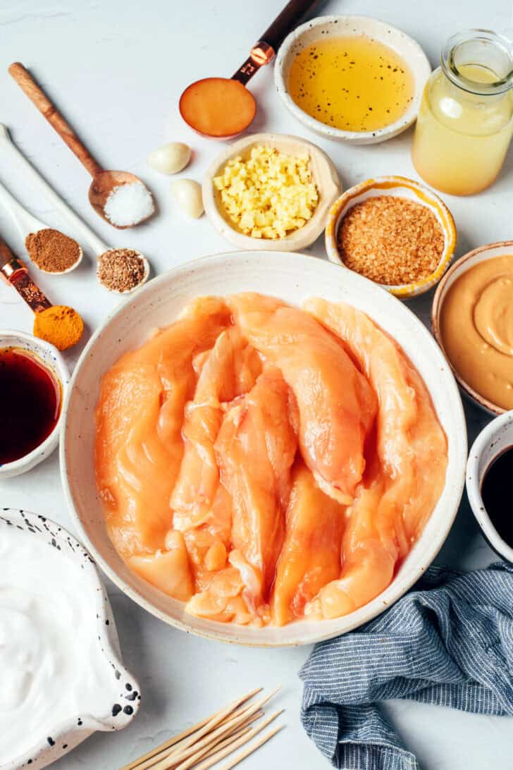 Pieces of chicken tender in a white bowl surrounded by small bowls with various recipe ingredients.