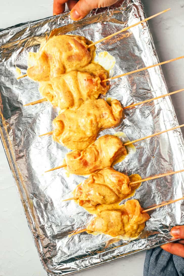 Hand holding a baking sheet covered with aluminum foil, holding 6 raw dipped chicken skewers.