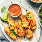 6 pieces of chicken satay on a white oval plate with slices of lime and a white ramekin of peanut sauce.