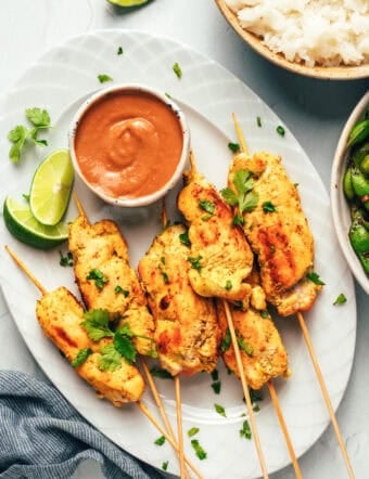 6 pieces of chicken satay on a white oval plate with slices of lime and a white ramekin of peanut sauce.