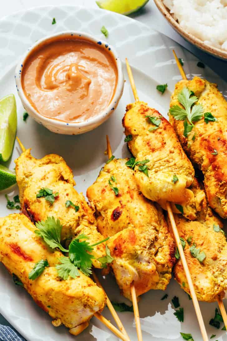 6 pieces of chicken satay on a white oval plate with slices of lime and a white ramekin of peanut sauce.