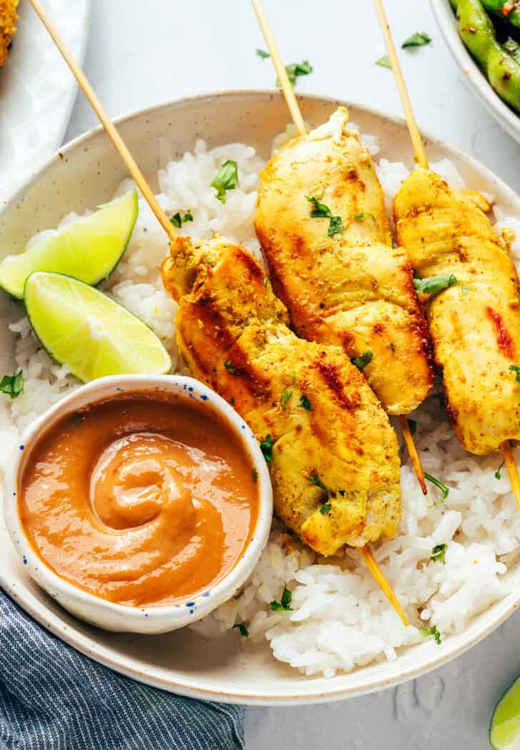 6 pieces of chicken satay on a white round plate with white rice, slices of lime and a white ramekin of peanut sauce.