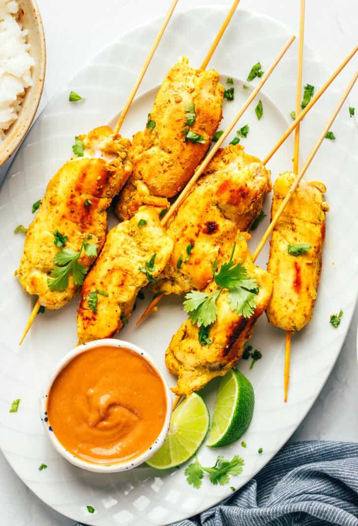 6 pieces of chicken satay on a white oval plate with slices of lime and a white ramekin of peanut sauce.