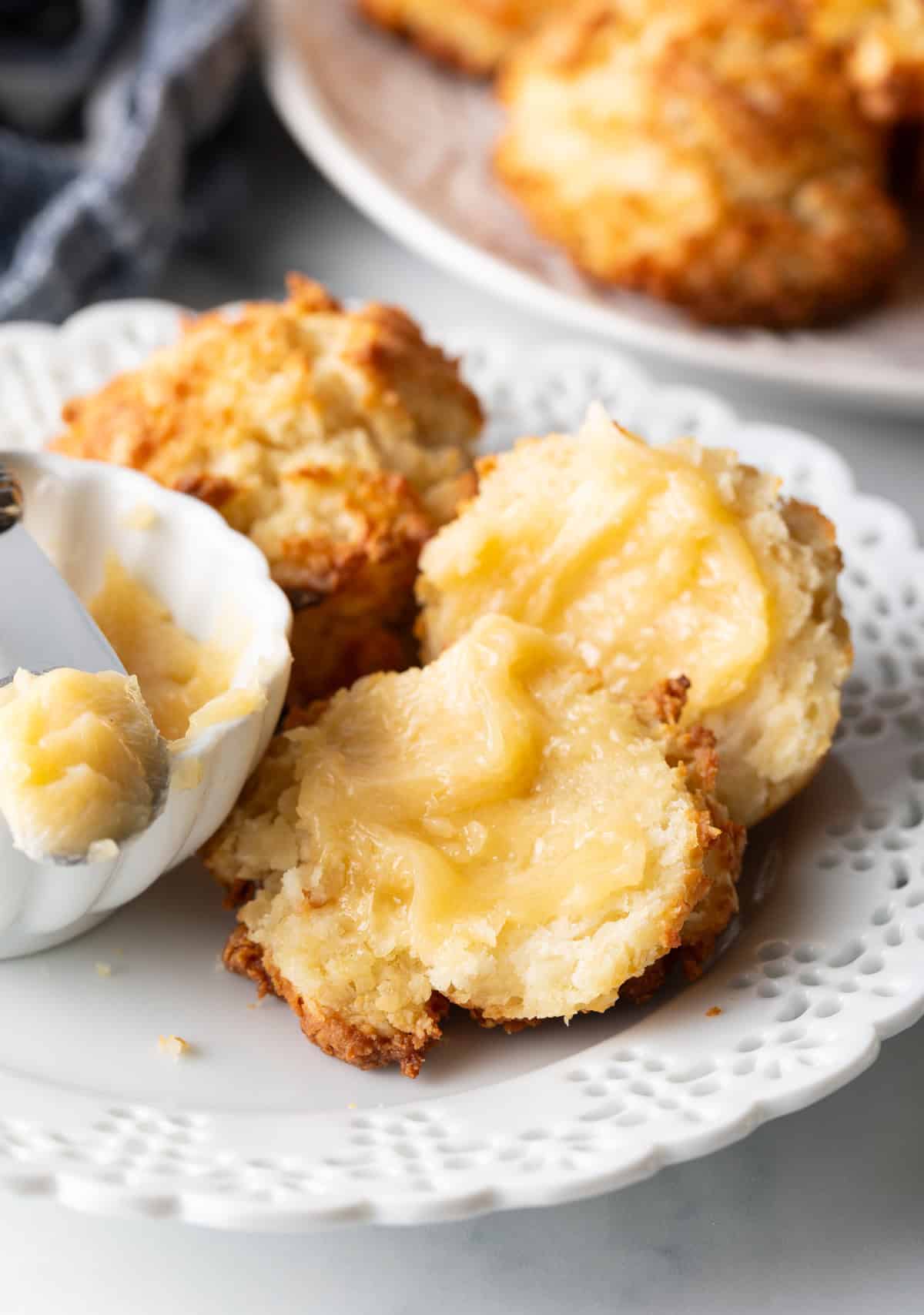 Close view of two buttermilk drop biscuits on a white plate, with a white ramekin of whipped honey butter and a butter knife showing a bit to camera. One biscuit is open with both sides buttered.