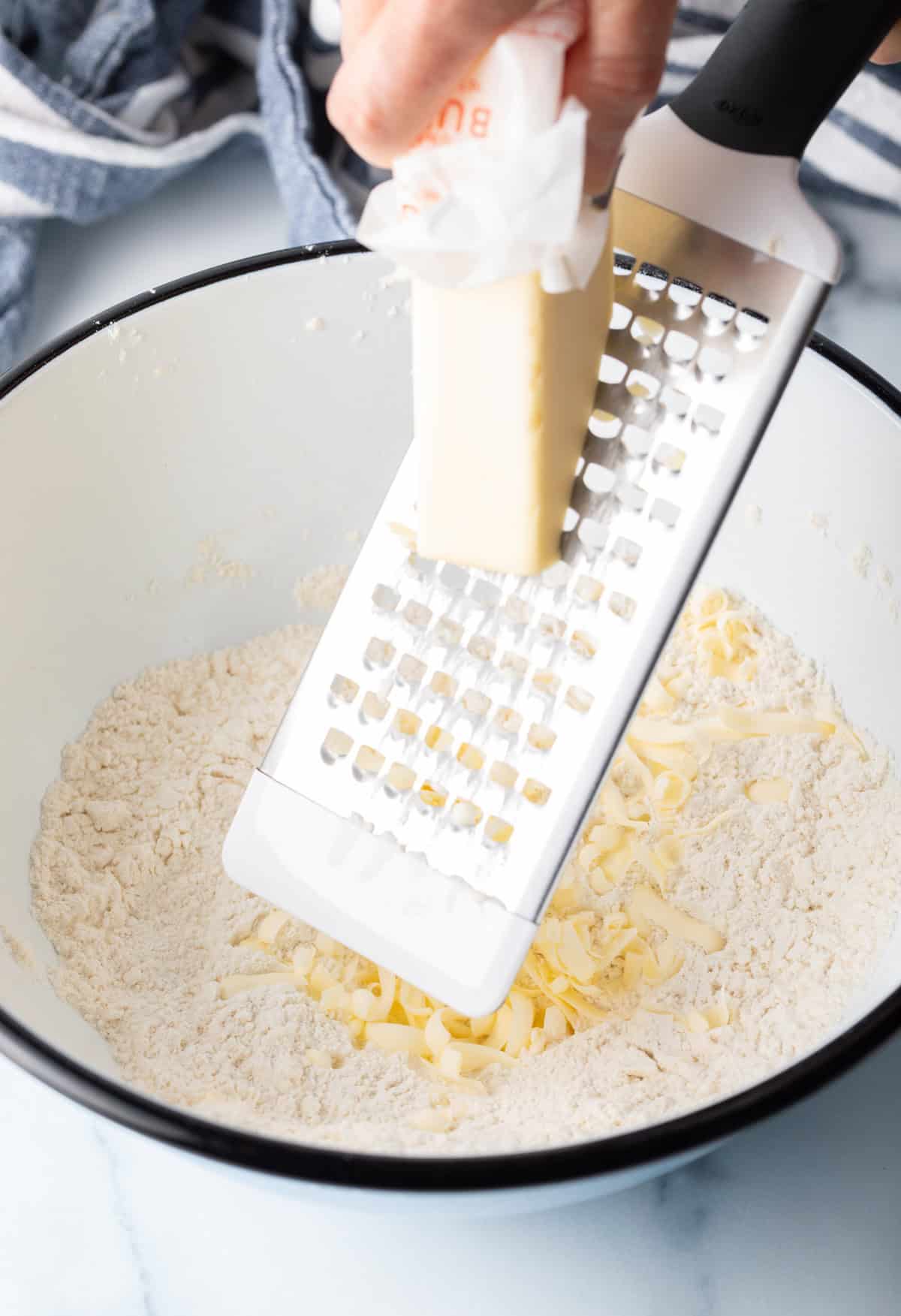 Using a cheese grater to grate butter into a white bowl with dry biscuit ingredients.