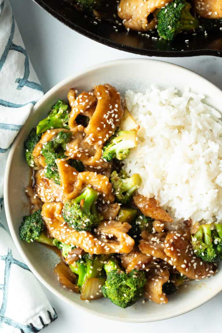 White shallow bowl with serving of Chinese chicken and broccoli with white rice.