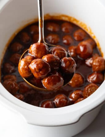White slow cooker filled with meatballs in bbq sauce. A ladle full of meatballs in being held to camera.