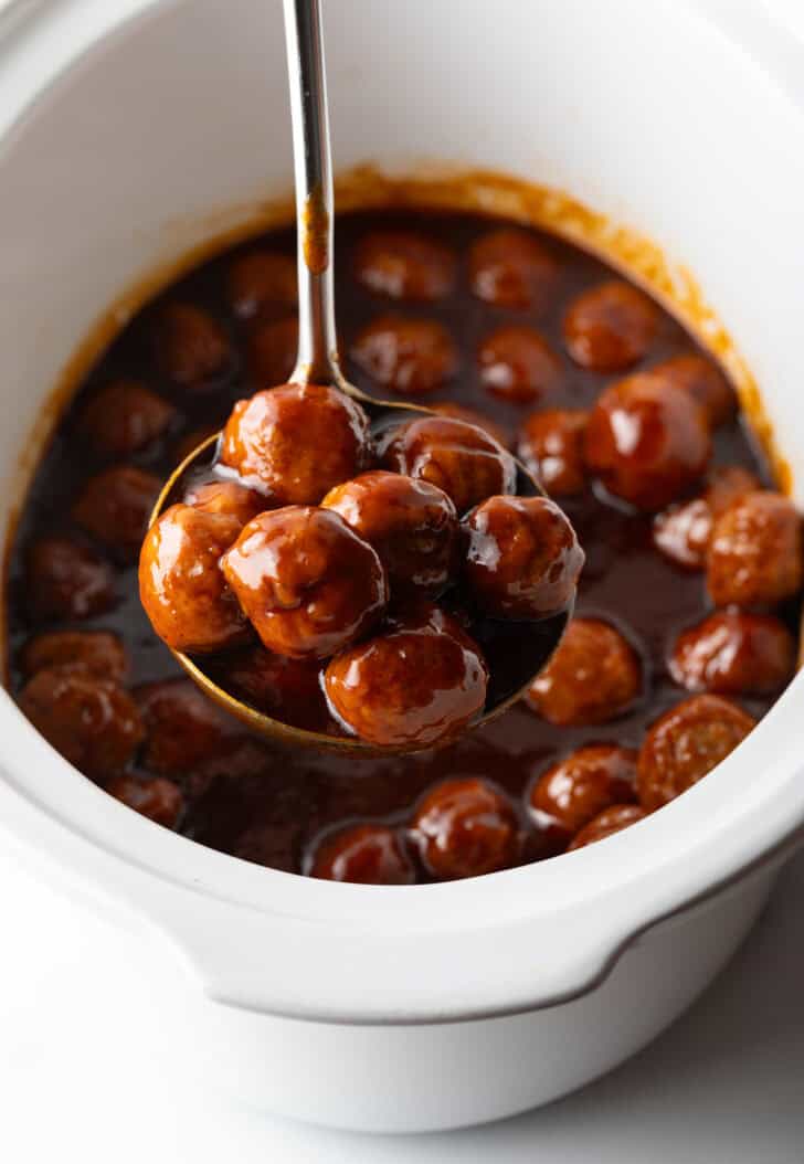 White slow cooker filled with meatballs in bbq sauce. A ladle full of meatballs in being held to camera.