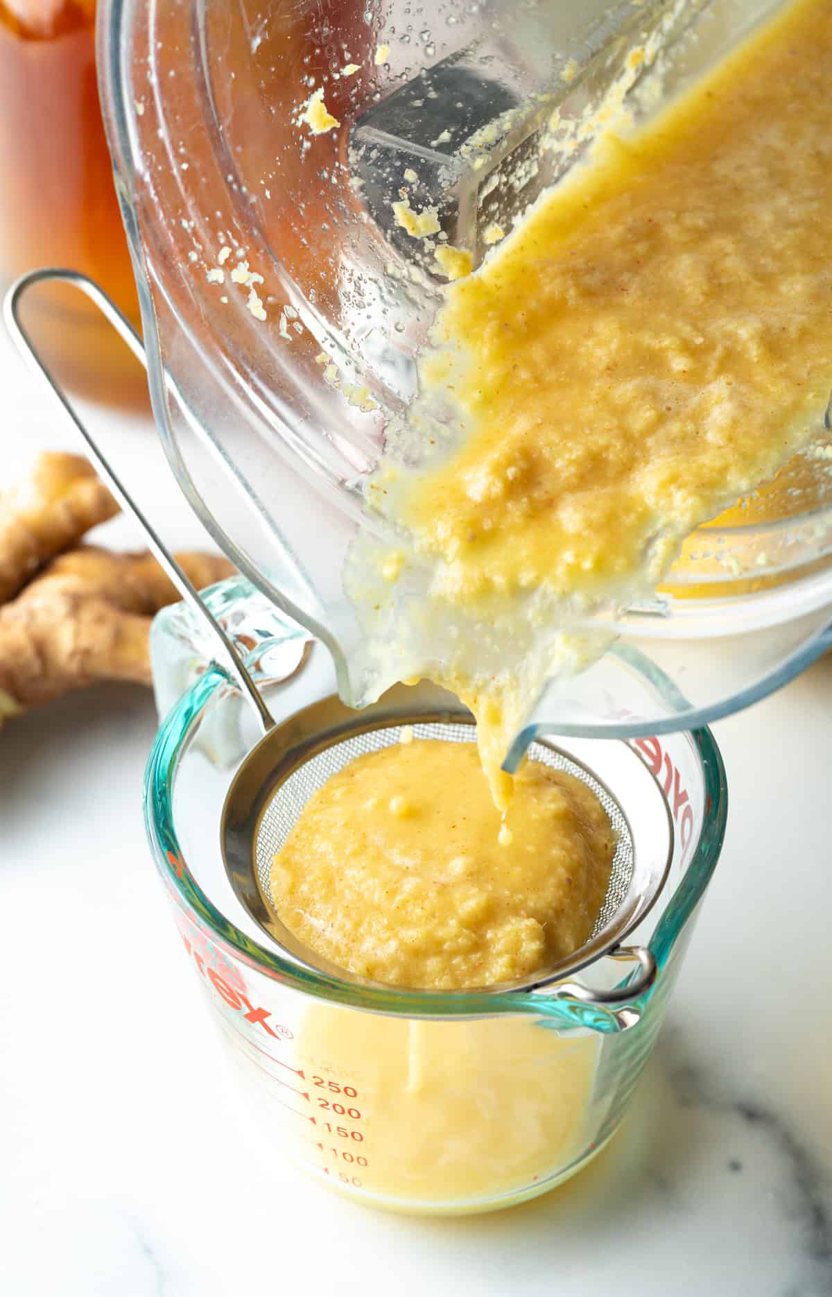Pouring pureed ginger from a food processor into a sieve set over a glass measuring pitcher.
