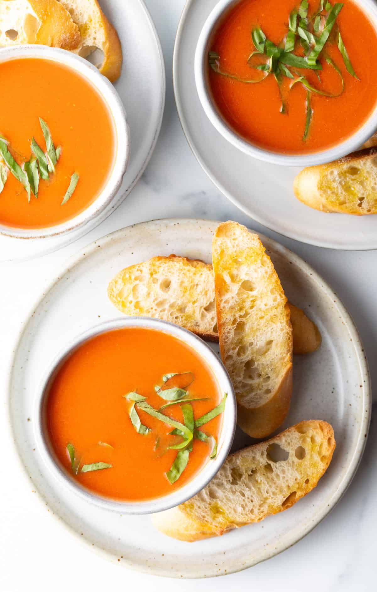 Top down view a bowl of tomato soup on a white plate, surrounded by three slices of baguette.
