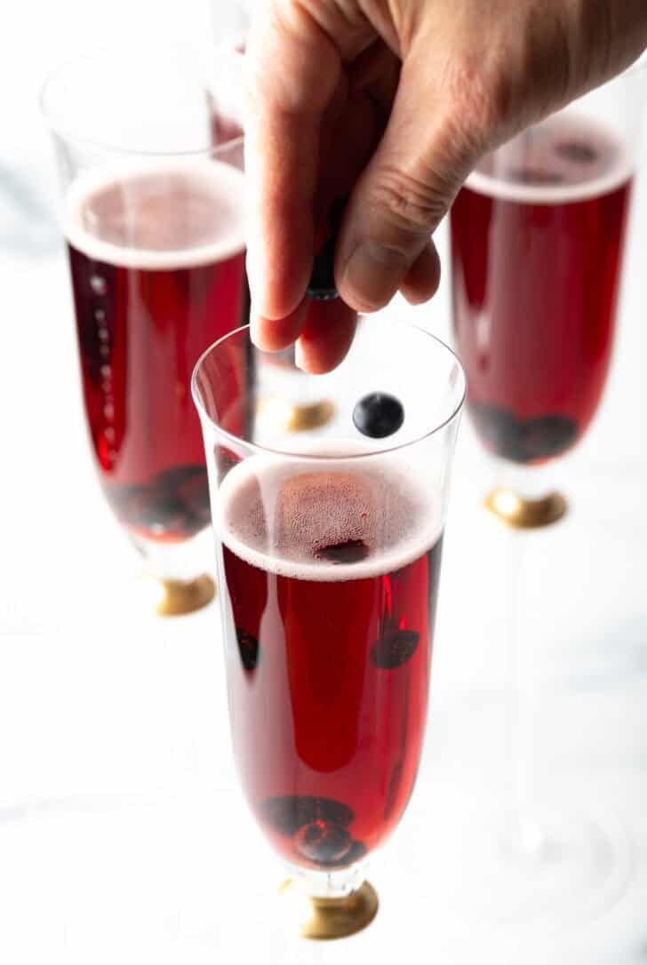 Hand sprinkling black currents into a champagne glass with red liquid.
