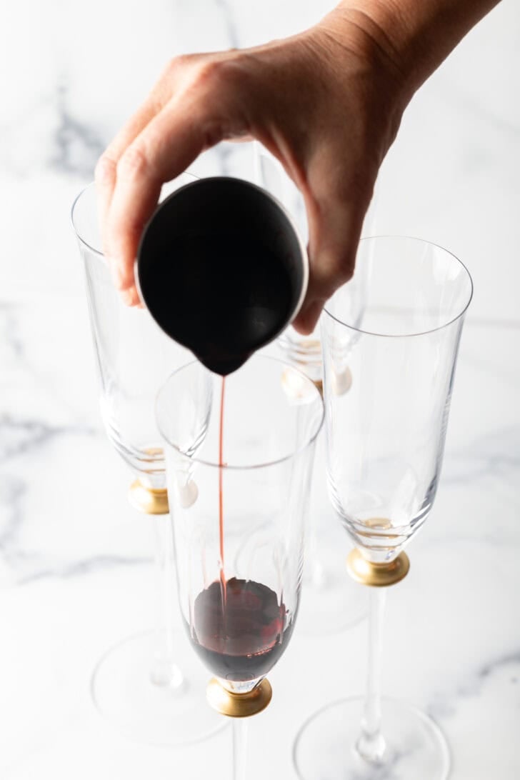 Hand pouring deep red liquid from a metal measuring cup into a fluted champagne glass.