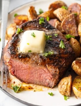 White plate with a reverse sear steak, cut in half to show the medium-rare meat inside. The steak is topped with chopped herbs and a pat of butter. Roasted yellow potato pieces are on the right side of the meat.