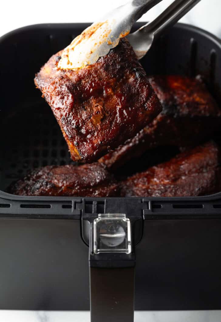 Tongs pulling a rib out of the air fryer and holding a cooked rib to camera.