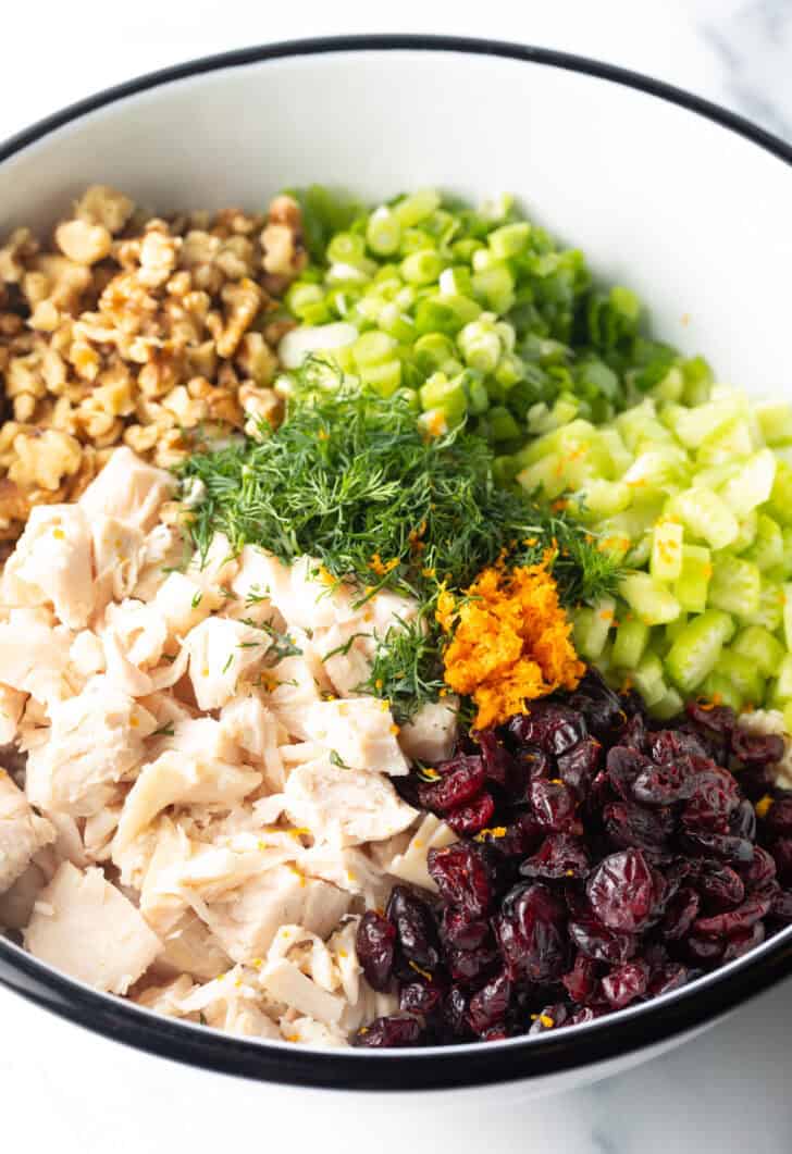White bowl with recipe ingredients in chopped piles: walnuts, celery, onions, dill, orange zest, cooked chicken, and cranberries.