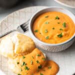 White plate with white bowl of red tomato gravy, next to a split open biscuit that's coated with the gravy and fresh chopped parsley.