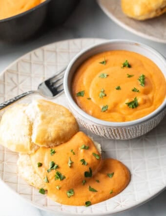 White plate with white bowl of red tomato gravy, next to a split open biscuit that's coated with the gravy and fresh chopped parsley.