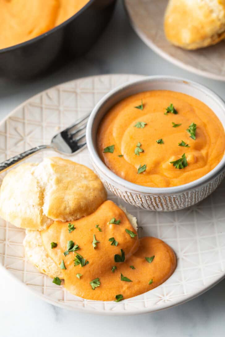 White plate with white bowl of red tomato gravy, next to a split open biscuit that's coated with the gravy and fresh chopped parsley.