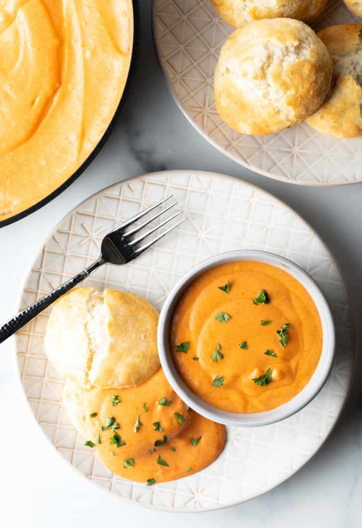 White plate with white bowl of red tomato gravy, next to a split open biscuit that's coated with the gravy and fresh chopped parsley.