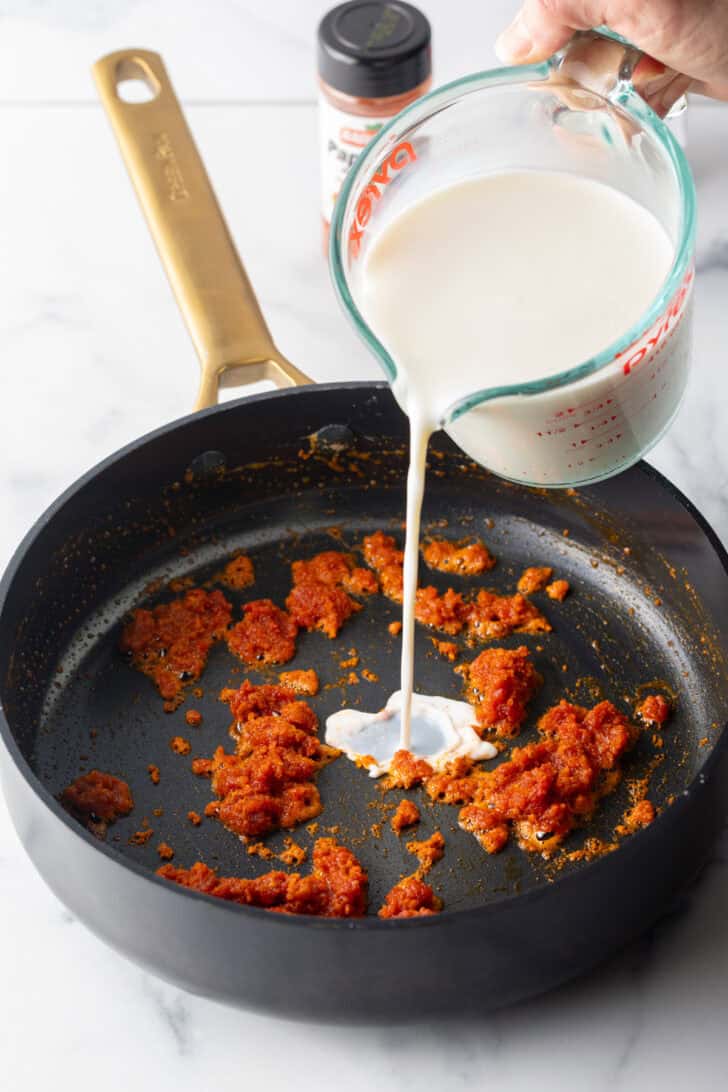 Pouring cream into a black skillet with tomato paste.