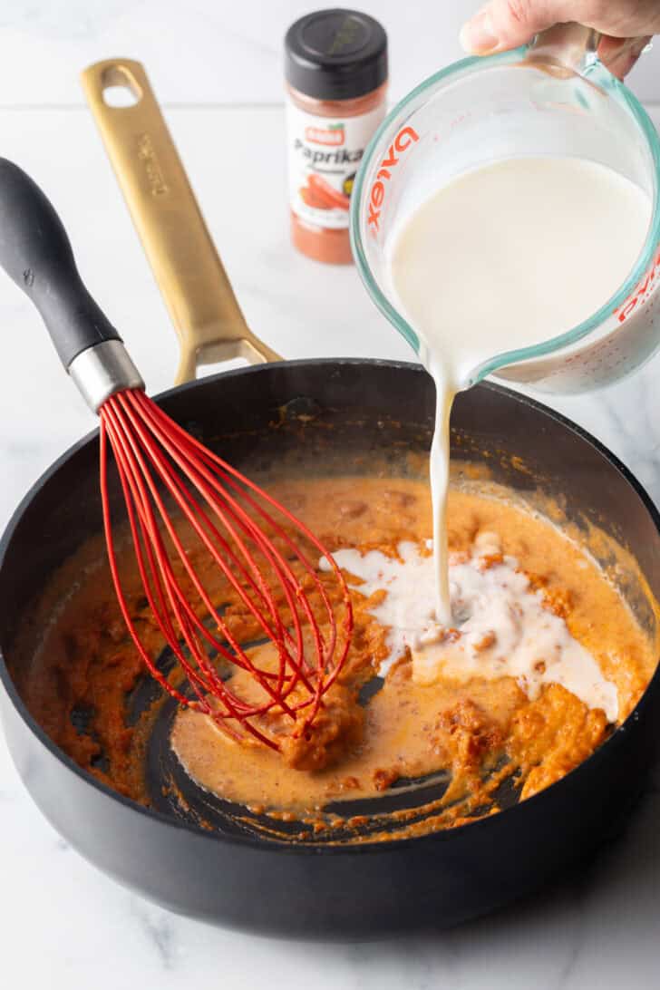 Pouring cream from a glass pitcher into a black skillet with tomato sauce and a red whisk.