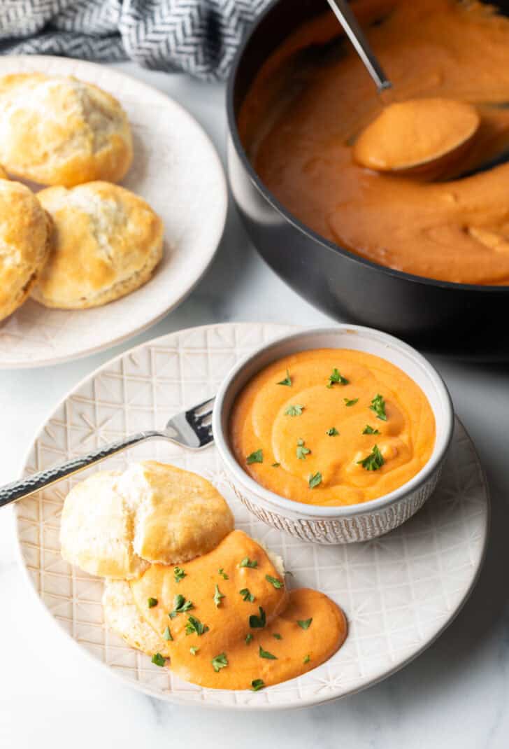 White plate with white bowl of red tomato gravy, next to a split open biscuit that's coated with the gravy and fresh chopped parsley.