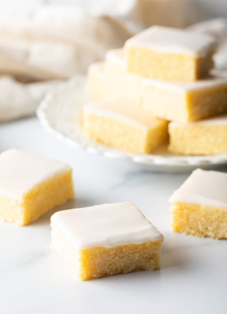 A lemon blondie towards camera, with a few bars and a stack of brownies on a white plate in the background.