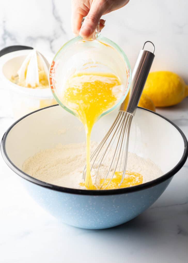 Pouring melted butter from a glass measuring cup into a large blue bowl with dry ingredients and whisk.