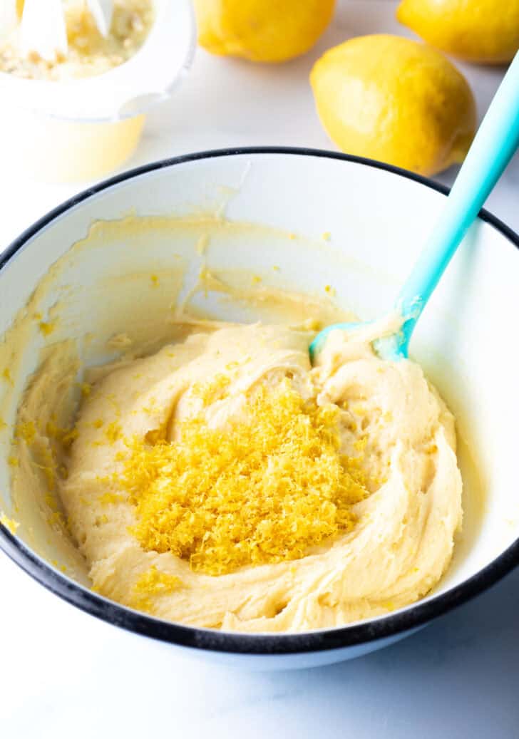 White mixing bowl with yellow brownie batter and lemon zest being stirred by a blue spatula.
